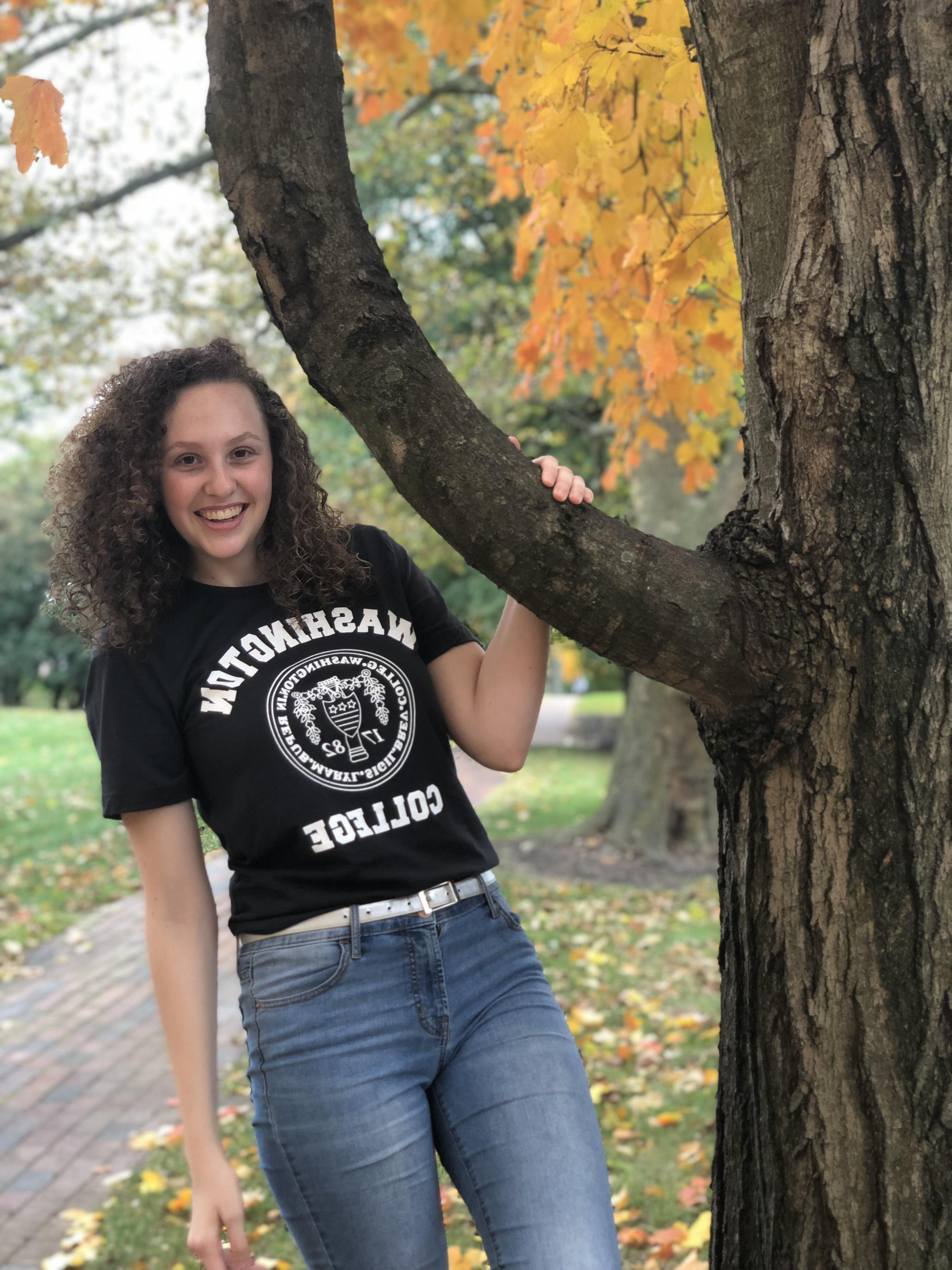 Carlee, 面带微笑，穿着印有九州娱乐官网标志的黑色t恤, blue jeans, and a white belt in front of fall foliage. 